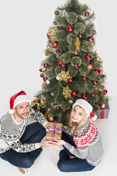 Високий кут зору пари в капелюхах Санта-Клауса, що тримає загорнутий подарунок біля ялинки ізольовані на білому — стокове фото