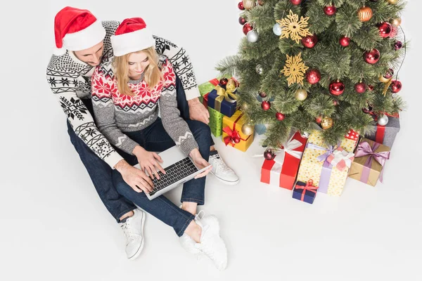 Vista aérea de la pareja en sombreros de santa claus usando el ordenador portátil cerca del árbol de Navidad aislado en blanco - foto de stock