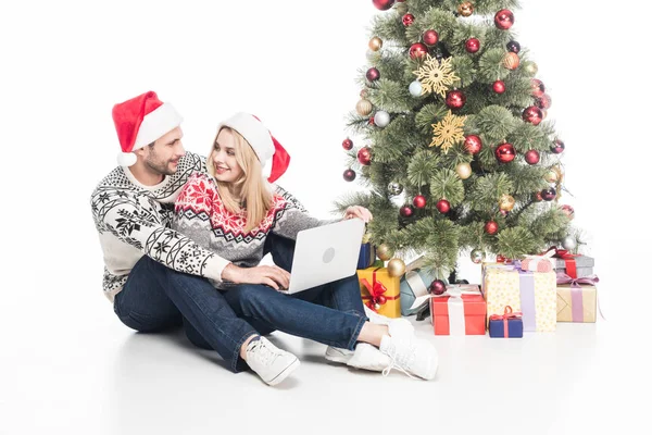 Sorrindo casal em santa claus chapéus com laptop perto de árvore de natal isolado no branco — Fotografia de Stock