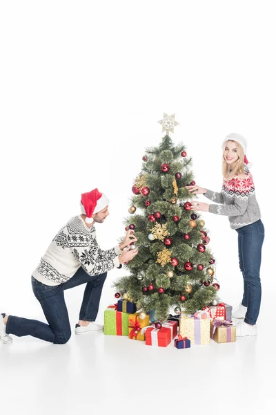 Junges Paar in Weihnachtsmannmützen schmückt gemeinsam Weihnachtsbaum isoliert auf weißem Grund — Stockfoto