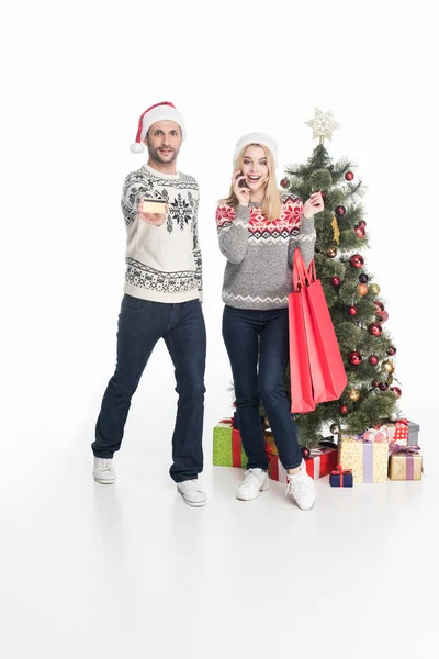 Casal em camisolas e chapéus de santa claus com sacos de compras perto da árvore de natal isolado em branco — Fotografia de Stock