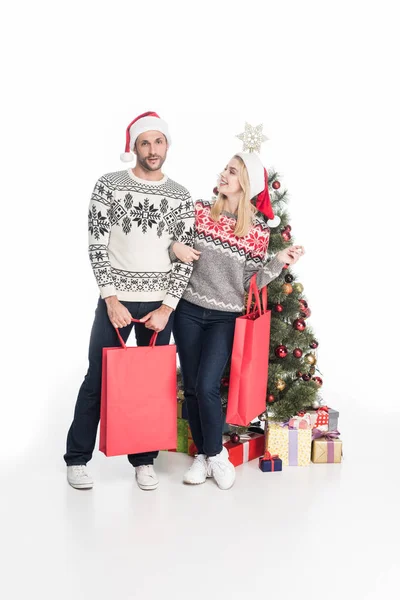 Pareja en suéteres y sombreros de santa claus con bolsas de compras cerca del árbol de Navidad aislado en blanco - foto de stock