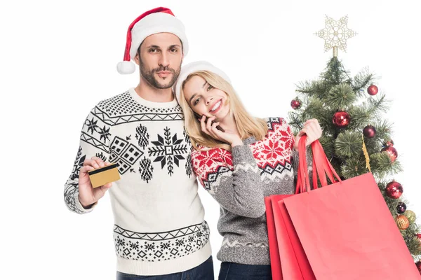 Jeune couple en pulls et chapeaux santa claus avec sacs à provisions et carte de crédit près de l'arbre de Noël isolé sur blanc — Photo de stock