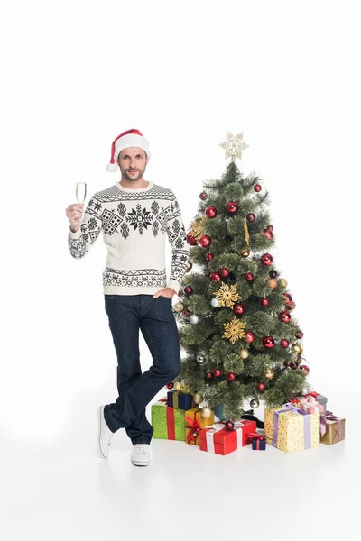 Hombre en suéter y sombrero de santa claus con copa de champán de pie cerca del árbol de Navidad aislado en blanco — Stock Photo