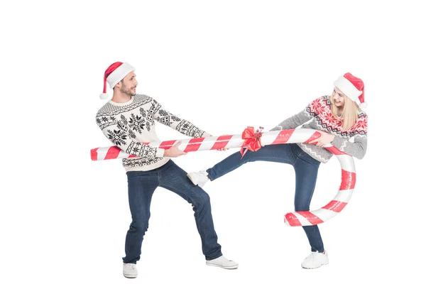 Young couple in santa claus hats and sweaters pulling over toy candy isolated on white — Stock Photo