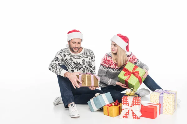 Pareja en suéteres y sombreros de santa claus con regalos envueltos sentados en el suelo aislados en blanco - foto de stock