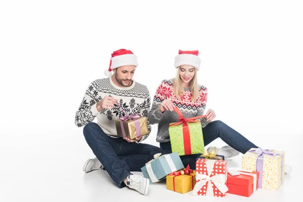 Couple in sweaters and santa claus hats unwrapping presents while sitting on floor isolated on white — Stock Photo