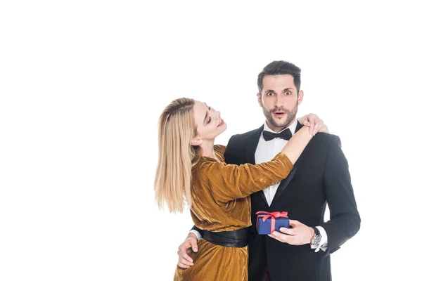 Attraktive Frau umarmt schockierten Freund im Anzug mit eingewickeltem Geschenk isoliert auf Weiß — Stockfoto