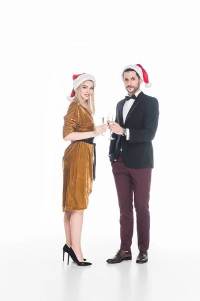 Vista lateral de la pareja de moda en sombreros de Santa Claus tintineo copas de champán aislado en blanco - foto de stock