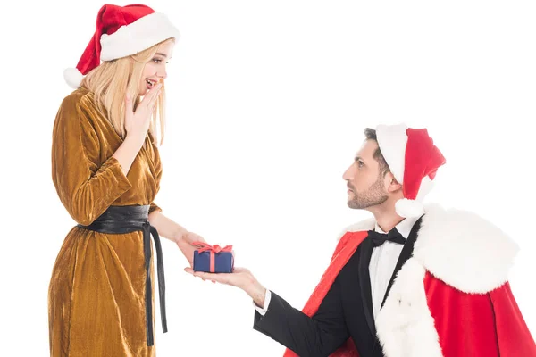 Man in santa claus costume presenting gift to surprised girlfriend in santa claus hat isolated on white — Stock Photo