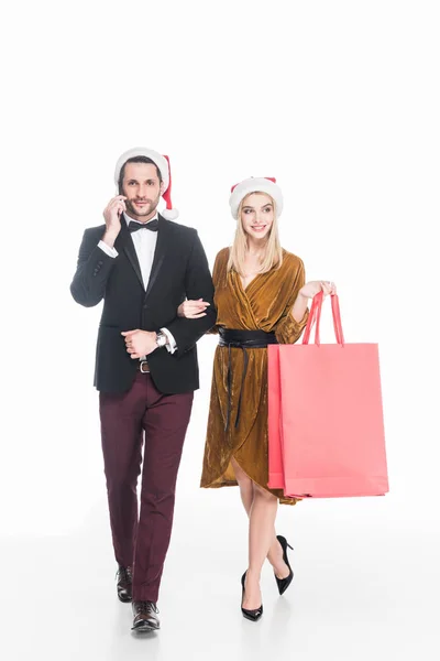 Atractivo hombre hablando en el teléfono inteligente cerca de novia en sombrero de Santa Claus con bolsas de compras aisladas en blanco - foto de stock