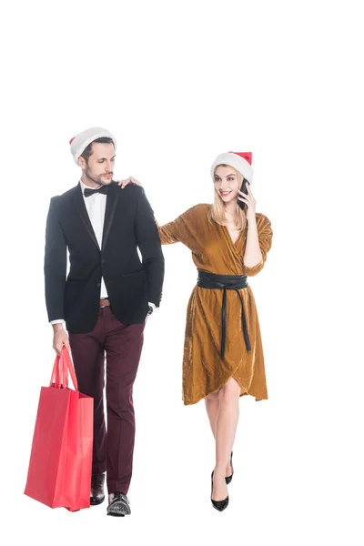 Sonriente mujer hablando en smartphone cerca de novio en santa claus sombrero con bolsas aisladas en blanco - foto de stock