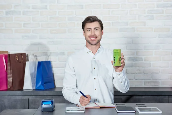 Vendedor masculino guapo sosteniendo teléfono inteligente con la mejor aplicación de compras y sonriendo a la cámara en la tienda - foto de stock