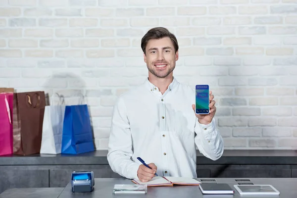Beau vendeur masculin tenant smartphone avec application de réservation et souriant à la caméra en magasin — Photo de stock