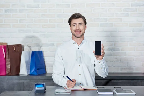 Apuesto joven vendedor sosteniendo teléfono inteligente con pantalla en blanco y sonriendo a la cámara en la tienda - foto de stock