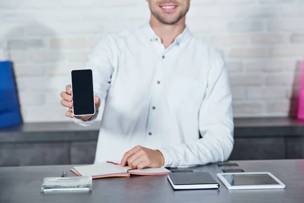 Recortado disparo de sonriente joven vendedor sosteniendo teléfono inteligente con pantalla en blanco en la tienda - foto de stock