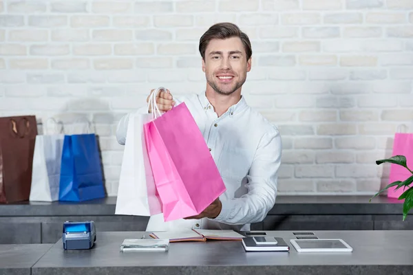 Apuesto joven vendedor sosteniendo bolsas de compras y sonriendo a la cámara en la tienda - foto de stock