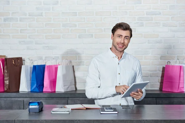 Красивый молодой продавец, использующий цифровой планшет и улыбающийся в магазине перед камерой — стоковое фото