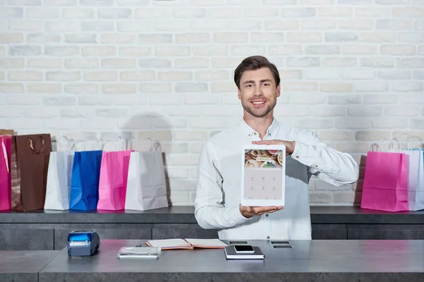 Schöner junger Verkäufer hält digitales Tablet mit Foursquare-Anwendung in der Hand und lächelt in die Kamera im Geschäft — Stockfoto