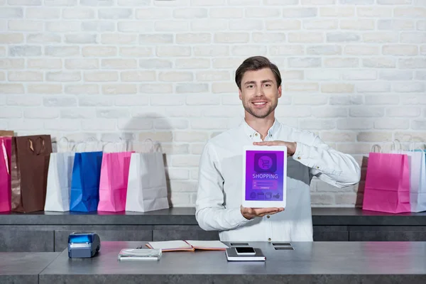 Apuesto joven vendedor sosteniendo tableta digital con aplicación de compras y sonriendo a la cámara en la tienda - foto de stock