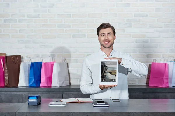 Apuesto joven vendedor sosteniendo tableta digital con solicitud de entradas y sonriendo a la cámara en la tienda - foto de stock