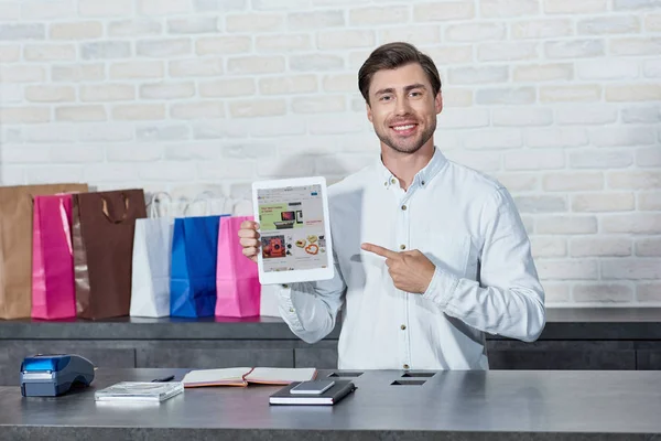 Beau jeune vendeur pointant vers tablette numérique avec application ebay et souriant à la caméra en magasin — Photo de stock