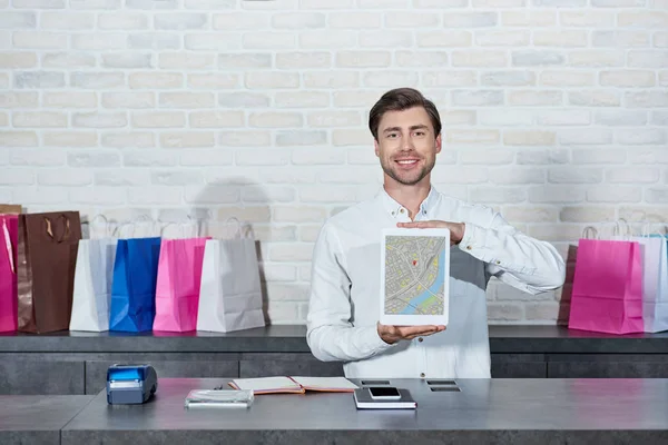 Schöner junger Verkäufer, der ein digitales Tablet mit Karte auf dem Bildschirm hält und im Geschäft in die Kamera lächelt — Stockfoto