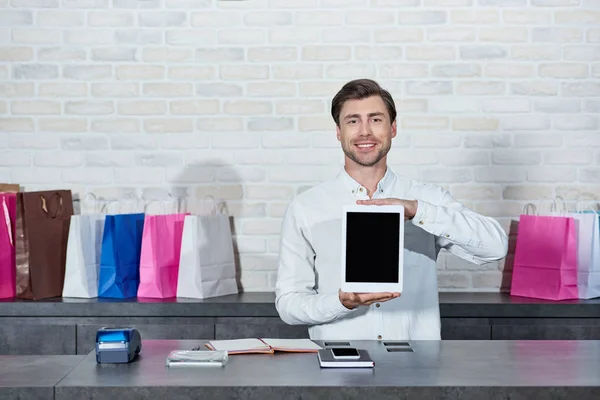 Schöner junger Verkäufer hält digitales Tablet mit leerem Bildschirm in der Hand und lächelt in die Kamera im Geschäft — Stockfoto