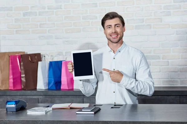 Schöner junger Verkäufer zeigt auf digitales Tablet mit leerem Bildschirm und lächelt in die Kamera im Geschäft — Stockfoto