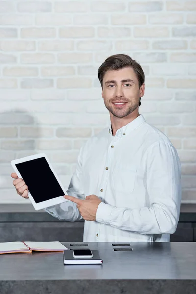 Schöner junger Verkäufer hält digitales Tablet mit leerem Bildschirm und lächelt in die Kamera im Geschäft — Stockfoto