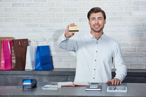 Handsome young salesman holding credit card and smiling at camera in shop — Stock Photo