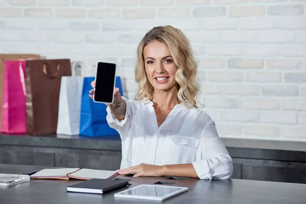 Hermoso joven vendedor sosteniendo teléfono inteligente con pantalla en blanco y sonriendo a la cámara en la tienda - foto de stock