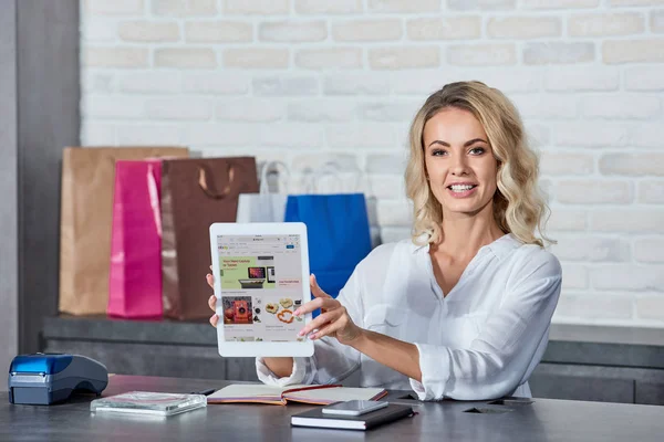 Hermosa mujer joven sosteniendo la tableta digital con la aplicación ebay y sonriendo a la cámara mientras trabaja en la tienda - foto de stock