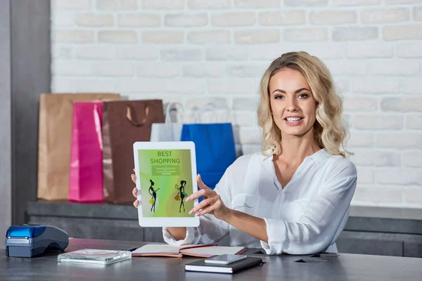 Schöne junge Frau hält ein digitales Tablet mit der besten Einkaufsanwendung in der Hand und lächelt in die Kamera, während sie im Geschäft arbeitet — Stockfoto
