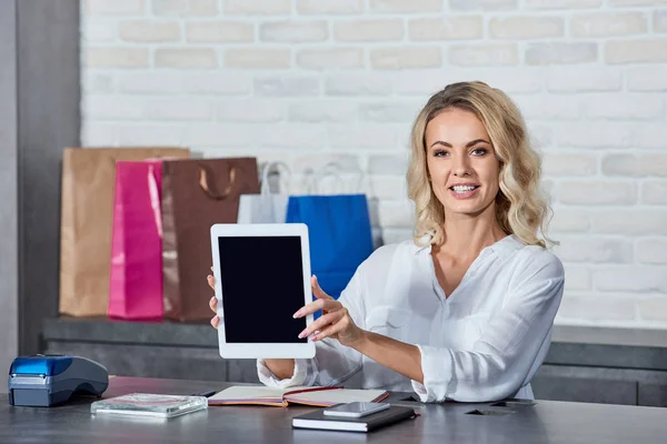 Hermosa mujer joven sosteniendo tableta digital con pantalla en blanco y sonriendo a la cámara mientras trabaja en la tienda - foto de stock