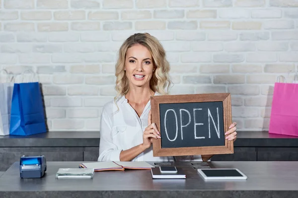 Glückliche junge Frau hält Schild offen und lächelt in die Kamera im Geschäft — Stockfoto
