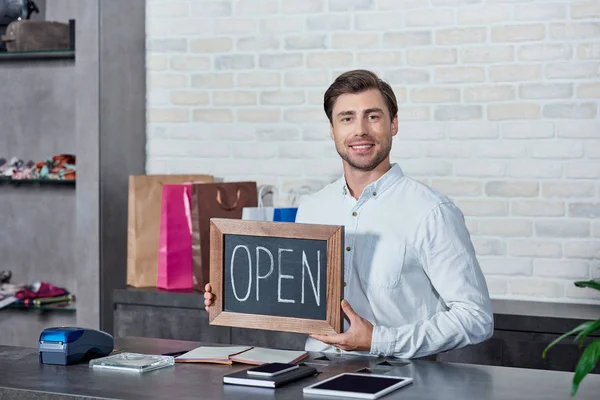 Schöner junger Verkäufer hält Schild offen und lächelt in die Kamera im Geschäft — Stockfoto