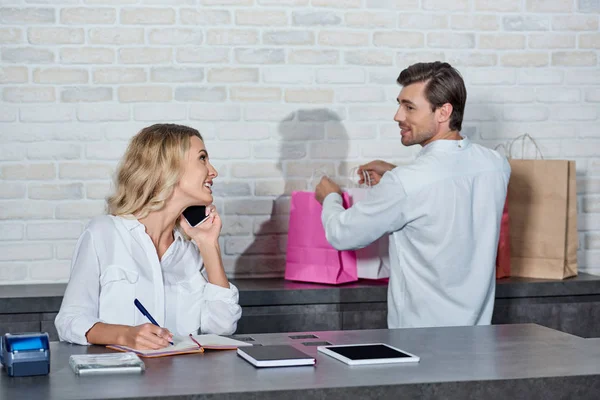 Junge Frau spricht mit Smartphone und blickt lächelnden Kollegen an, der Einkaufstüten im Laden hält — Stockfoto