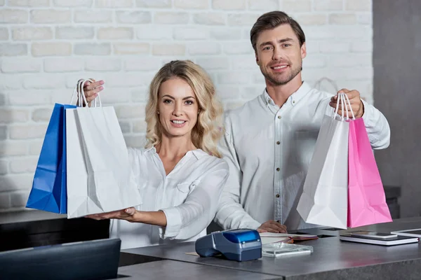 Happy young shop workers holding paper bags and smiling at camera in store — Stock Photo