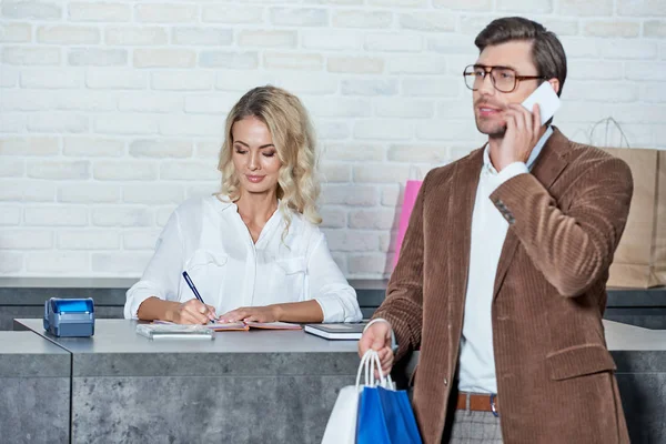 Uomo sorridente che tiene borse della spesa e parla con lo smartphone mentre il venditore femminile prende appunti nel negozio — Foto stock