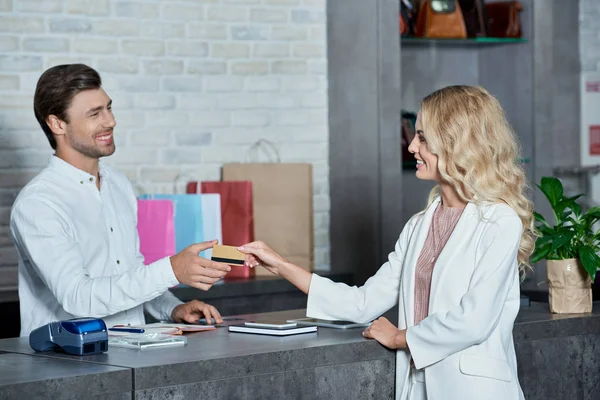 Beautiful young female customer giving credit card to smiling salesman in shop — Stock Photo