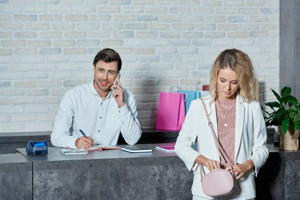 Smiling seller talking by smartphone and female customer looking into bag in store — Stock Photo