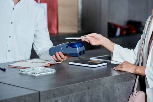 Tiro cortado de trabalhador segurando terminal de pagamento e mulher pagando com smartphone na loja — Fotografia de Stock