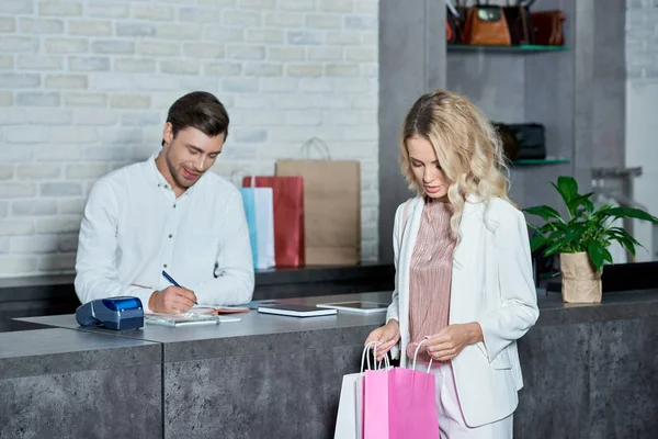 Giovane donna che guarda nelle borse della spesa mentre il venditore prende appunti in negozio — Foto stock