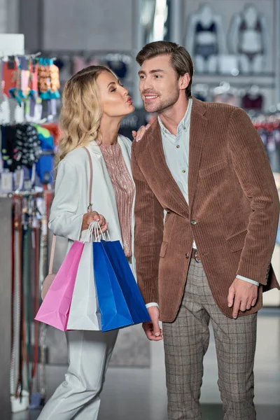 Щаслива жінка тримає сумки для покупок і цілує усміхненого чоловіка в магазині — стокове фото