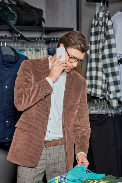 Joven en gafas de vista hablando por teléfono inteligente y mirando ropa con estilo en la tienda - foto de stock