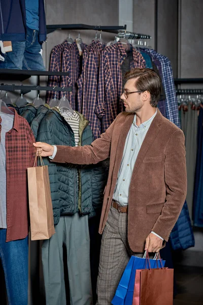 Handsome man holding shopping bags and choosing clothes in store — Stock Photo