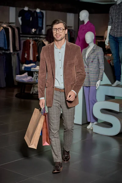 Bell'uomo con gli occhiali che tiene le borse della spesa e sorride alla macchina fotografica nel negozio di moda — Foto stock