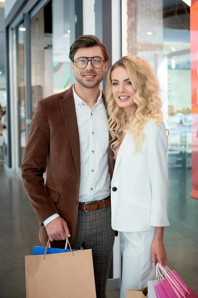 Feliz joven pareja con bolsas de compras sonriendo a la cámara en el centro comercial - foto de stock