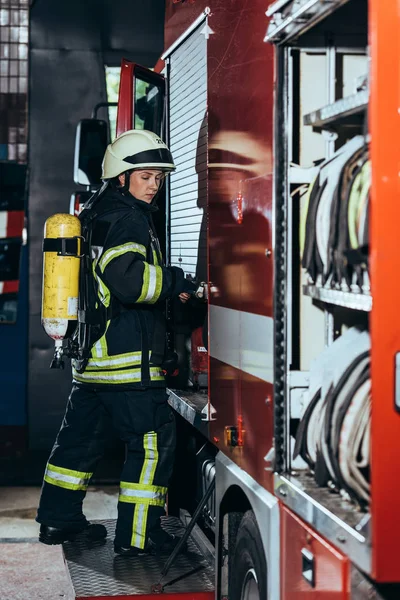 Side view of female firefighter with fire extinguisher on back closing truck at fire station — Stock Photo